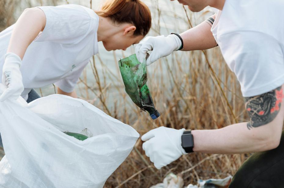Woman and man cleaning up. She is holding rubbish bag while man wearing gloves throws an empty plastic bottle in